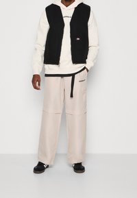 Černá bezrukávová vesta přes krémovou mikinu, doplněná světle béžovými kalhotami s širokými nohavicemi, které mají boční kapsu a černé akcenty. Černé sneakers.