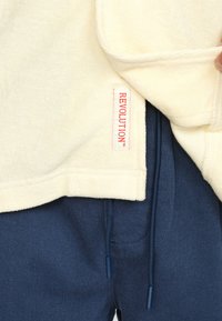Cremfarbener Oberteil mit einem roten "REVOLUTION"-Label, kombiniert mit navyblauen Jogginghosen mit glatter Oberfläche.