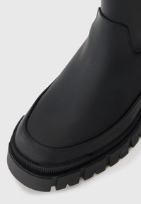 Schwarzer Gummistiefel mit einer groben Profilsohle, glatter Textur und runder Form. Verfügt über eine dezente Nahtdetails entlang der oberen Kante.