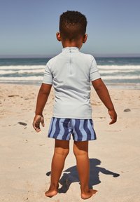 Maglietta da rash guard a maniche corte di colore azzurro con una cerniera sul retro, abbinata a pantaloncini da bagno a righe blu navy e azzurro, in piedi su una spiaggia sabbiosa.