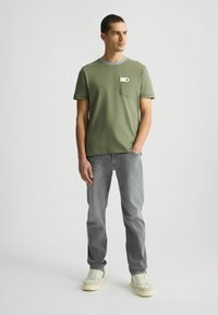 Camiseta verde oliva con cuello redondo, manga corta y un bolsillo en el pecho. Combinada con jeans gris claro y zapatillas de color crema.
