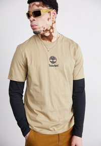 Trumpas, šviesiai rudos spalvos marškinėliai su juodomis ilgomis rankovėmis, sluoksniuotomis po jais, su Timberland logotipu ant krūtinės; papildyta auksiniais akinių nuo saulės.