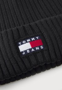 Gorro de punto acanalado negro con una etiqueta de tela que presenta un logotipo en azul, rojo y blanco que dice "TOMMY JEANS". Textura suave y diseño ajustado.