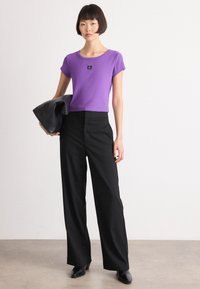 Purple ribbed t-shirt with a small black logo, paired with loose black trousers and a black leather bag, set against a light background.