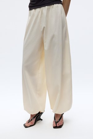 Person wearing loose, wide-legged cream pants paired with black heeled thong sandals against a plain background.