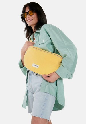 Femme souriante portant des lunettes de soleil à verres jaunes, une chemise oversize vert clair, un short en jean, tenant un grand sac zippé jaune en bandoulière sur la poitrine.