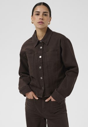 Femme aux cheveux foncés et boucles d’oreilles en or en forme d’anneaux, portant une veste en jean marron foncé boutonnée et un pantalon assorti, les mains dans les poches.