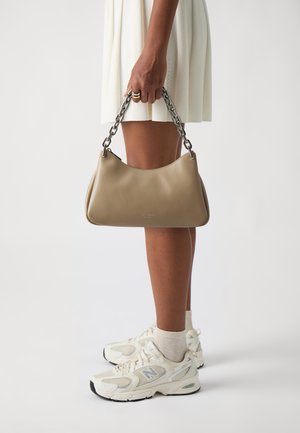 Bolso de cuero color marrón claro con una silueta curvada, que cuenta con un asa de cadena plateada. Llevado con un vestido blanco con plisados y zapatillas color crema.