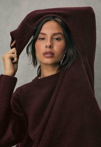 Bordeaux sweater med en struktureret, fuzzy finish; rund hals og ribbede manchetter; modelen poserer med den ene arm hævet, hvilket fremhæver tøjets fald.