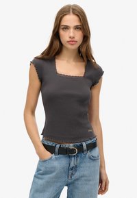 Grauer gerippter Oberteil mit kurzen Ärmeln, quadratischem Ausschnitt und gewellten Kanten. Kombiniert mit hellblauen, hoch taillierten Jeans und einem schwarzen Gürtel.
