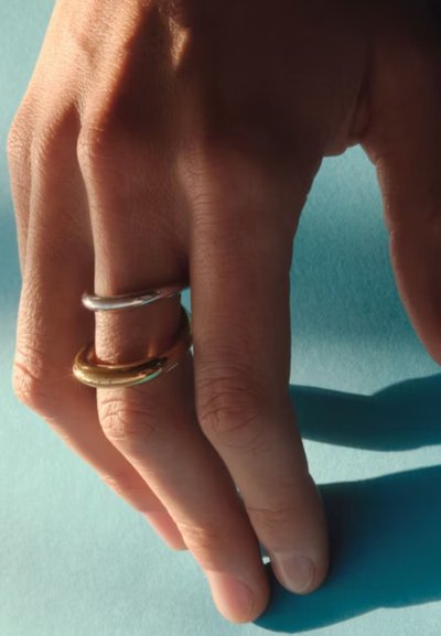Hand mit zwei dicken, glänzenden Ringen, einem silbernen und einem goldenen, die einen Schatten auf einen hellblauen Hintergrund werfen.