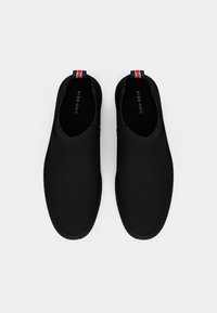 Mustat liukkaat kengät, joissa on sileä pinta, pyöreä siluetti ja erottuva punavalkoisen ja sinisen raidallisen vetoketjun nyöri kantapäässä.