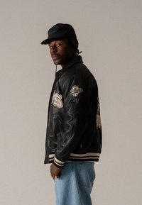 Black leather bomber jacket with embroidered patches, ribbed cuffs and hem, paired with light blue denim pants and a black bucket hat.
