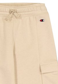 Beige Jogginghose mit einem elastischen Bund und Zugband, seitlichen Taschen und einem kleinen Logopatch am oberen linken Bein.
