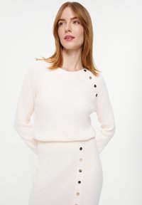 Hellrosa gerippter Strickpullover mit rundem Halsausschnitt und dekorativen Knöpfen an der Schulter, kombiniert mit einem passenden Rock, der seitliche Knöpfe aufweist.