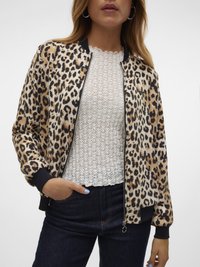 Femme portant une veste zippée à imprimé léopard beige et noir sur un haut en dentelle blanche et un jeans bleu foncé, avec une main dans la poche.