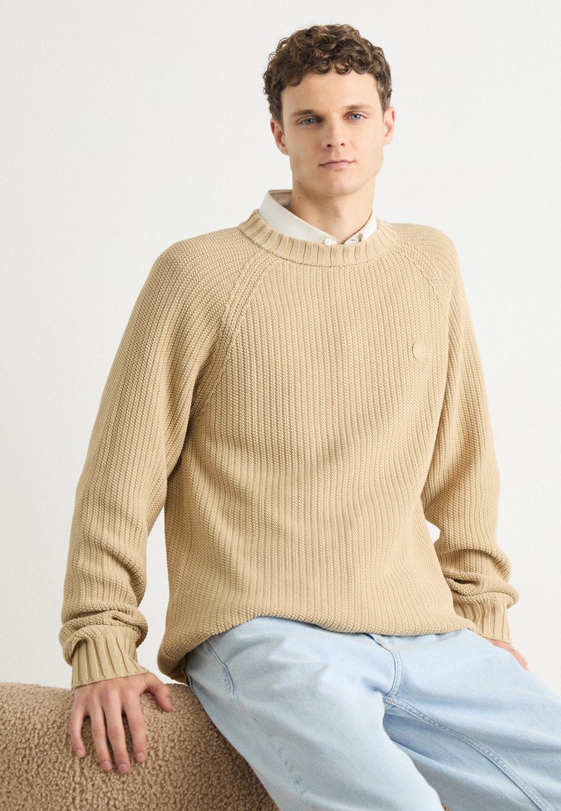 Beige ribbed knit sweater with a round neckline and cuffs, paired with light blue jeans. Features subtle logo detail on the chest.