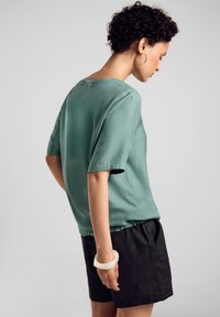 Satin green short-sleeve blouse with a round neckline, paired with black shorts. A translucent bangle adorns the wrist. Simple and relaxed design.