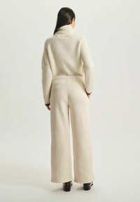 Pull en crème épais avec texture côtelée et col cheminée, associé à un pantalon large beige. Tissu lisse et contraste de couleurs subtil.