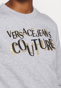 Close-up of light gray sweatshirt with black and gold glitter text reading "Versace Jeans Couture" on chest.