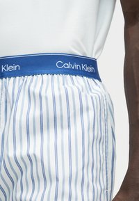 Ζώνη μέσης με μπλε λογότυπο Calvin Klein σε λευκά και μπλε κάθετα ριγωτά boxer, φορεμένα με λευκό πουκάμισο.