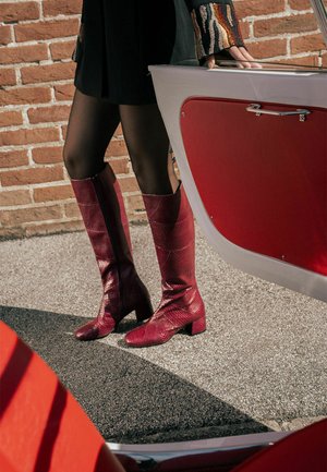 Bottes en cuir rouges montantes jusqu'aux genoux avec une finition texturée et une fermeture éclair latérale, associées à des collants noirs et une courte robe noire, posées à côté d'un bateau rouge.