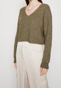 Pull court vert olive avec col en V, motif en maille texturée et manches longues, associé à un pantalon taille haute beige clair.