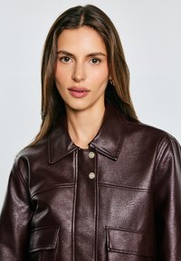 Burgunderfarbene Kunstlederjacke mit Kragen, zwei Vordertaschen und Druckknopfverschluss; glatte Textur mit leichtem Glanz.