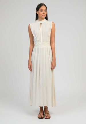 Jeune femme aux longs cheveux foncés portant une robe maxi blanche plissée sans manches et des sandales marron à brides, debout devant un fond uni.