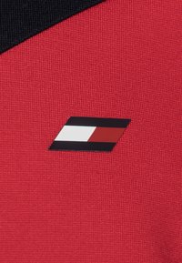 Ein roter Strukturstoff zeigt ein schwarz-weißes Logo mit einem roten Akzent, das sich auf der linken Seite, in der Nähe der Schulter, befindet.