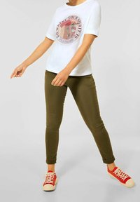 Biała koszulka z różowym nadrukiem graficznym, oliwkowe wąskie dżinsy i czerwone trampki z białymi podeszwami. Gładki materiał, swobodny krój, podwinięte mankiety.