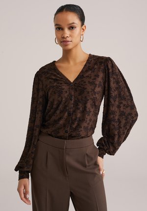Blouse marron à motif floral avec décolleté en V, fermeture par boutons, et manches longues bouffantes. Portée avec un pantalon marron assorti et ajusté.