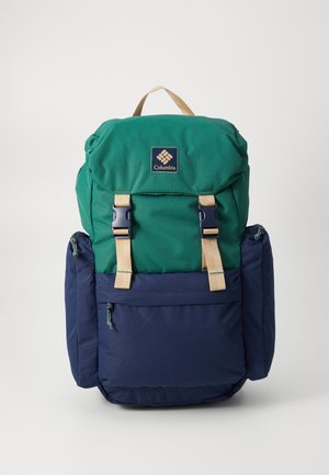 Sac à dos vert et bleu marine en tissu texturé, avec deux poches avant, des sangles beige et une étiquette de logo à l'avant. Comprend des compartiments zippés.