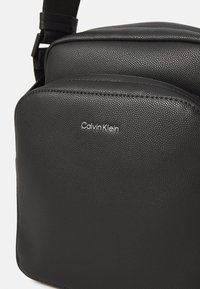 Borsa a tracolla in ecopelle nera con superficie texturizzata, scomparti con zip e logo Calvin Klein in argento. Inclusa una tracolla regolabile.