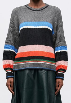 Personne portant un pull tricoté avec de larges rayures horizontales multicolores et une jupe en cuir vert foncé, mains visibles sur les côtés.
