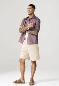 Kortærmet, lyserød skjorte med knapper, hvid t-shirt under, beige shorts, brune sandaler, stående pose med krydsede arme.