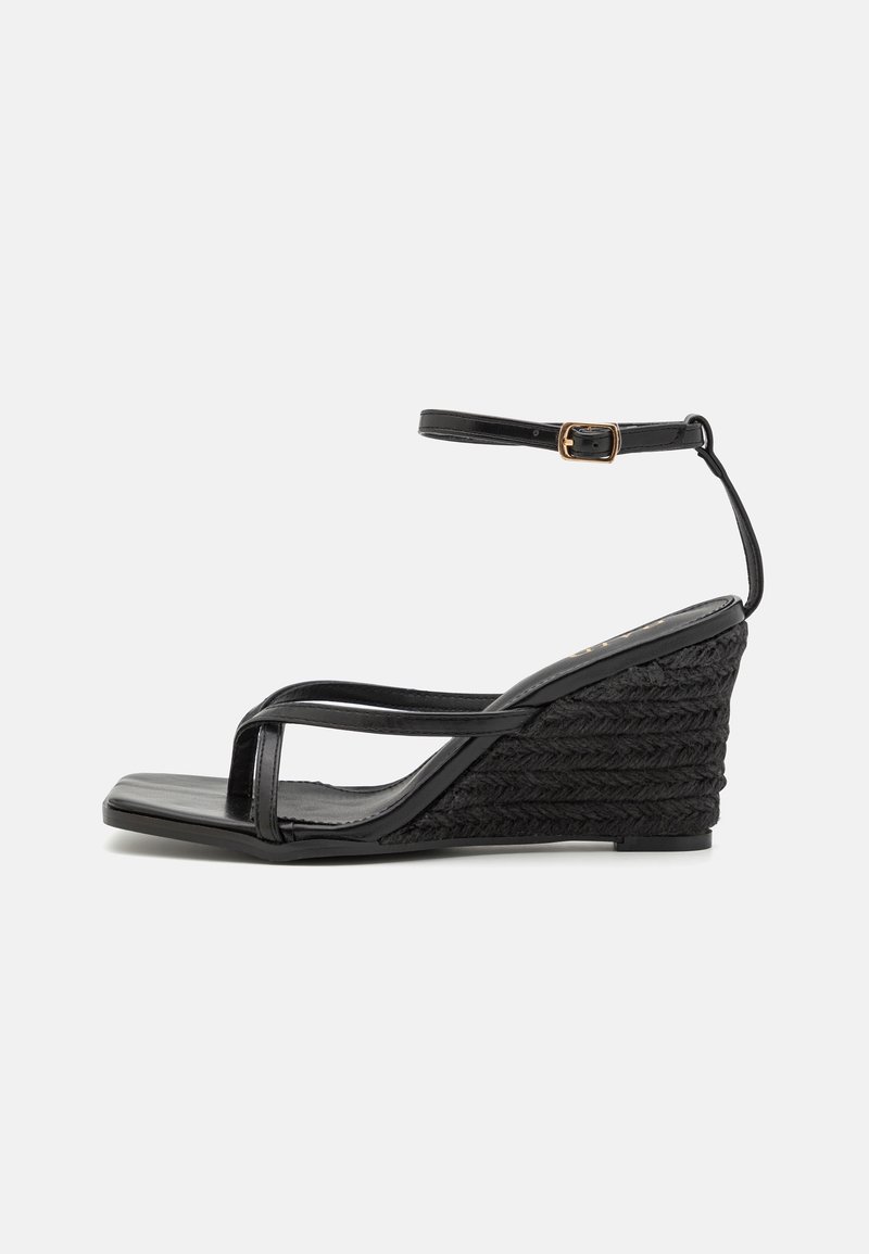Sandale compensée noire avec talon à texture tissée, fine bride sur le devant et bride de cheville ajustable avec boucle dorée, vue de côté gauche sur un fond uni.