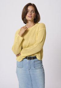 Suéter amarillo de punto con un ajuste holgado, que presenta una textura de canalé y mangas largas. Combinado con una falda de mezclilla azul claro y un cinturón negro.