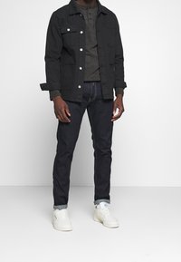 Schwarze Jacke mit silbernen Knöpfen über einem dunkelgrauen Henley-Shirt, kombiniert mit dunkelblauen Jeans und weißen Sneakers. Einfaches Design und bequeme Passform.