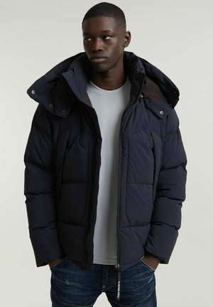 Navy padded jacket with a high collar and detachable hood, featuring zip closure and visible stitching details; worn over a light gray shirt.