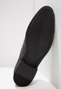 Semelle de chaussure habillée en cuir noir, face à l'appareil photo, appuyée contre un mur d'angle blanc sur une surface blanche.