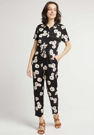 Combinaison noire à fleurs avec manches courtes, taille élastique et poches latérales. Présente de grandes fleurs blanches et une coupe décontractée. Talons marron.