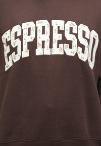 Sudadera marrón con la palabra "ESPRESSO" en grandes letras blancas y pequeños detalles dorados. Textura suave, corte holgado y puños acanalados.