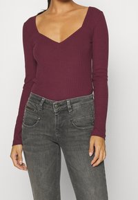 Frau, die ein rippendes, langärmeliges, dunkelrotes V-Ausschnitt-Oberteil trägt, kombiniert mit verwaschenen schwarzen Jeans, die mit Reißverschlüssen und Nieten verziert sind.