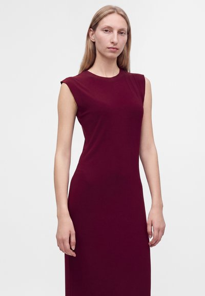 Femme aux longs cheveux blonds portant une robe rouge foncé sans manches, debout devant un fond clair uni.