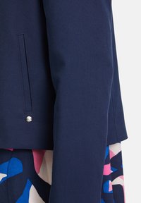 Nahaufnahme des Ärmels und der Taschendetails einer dunkelblauen Jacke über einem mehrfarbigen Rock mit abstrakten Mustern in Blau, Pink und Weiß.