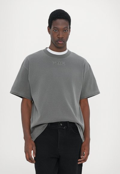 T-shirt gris à manches courtes avec un logo texturé sur la poitrine, encolure ronde et coupe décontractée, associé à un jean noir.