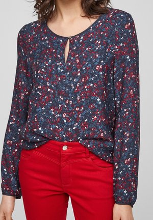 Vrouw draagt een marineblauwe blouse met rood-wit bloemmotief en rode broek, blouse losjes in de taille gestopt.