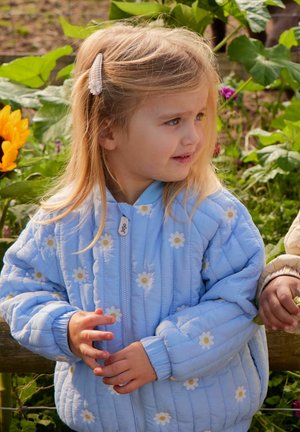 Fiatal lány szőke hajjal és virágos hajcsattal, világoskék margarétamintás steppelt dzsekit visel, szabadtéren áll, zöld növényzet és napraforgók közelében.