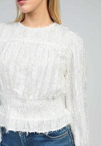 Femme portant un blouse blanche texturée à manches longues avec une taille smockée et un jean bleu, vue des épaules aux hanches.